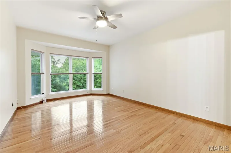 Bright room featuring a large bay window, wood-finish flooring, a ceiling fan with integrated lighting, and neutral wall tones — 3515 Truman Terrace Drive #65, St Charles, MO 63301