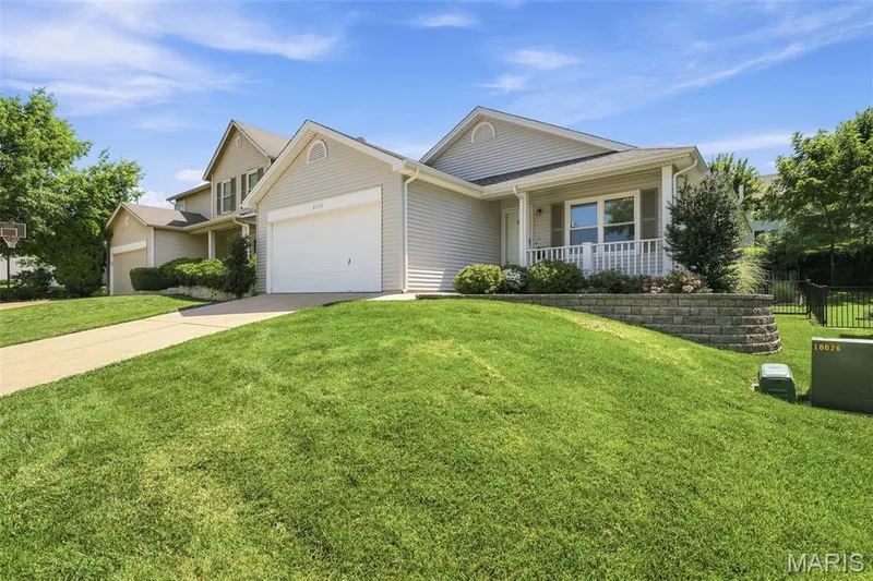 Light-colored siding exterior featuring an attached garage with a white door, a covered front porch with white railing, a stone retaining wall, and a green lawn — 2510 Balaji Court, Wildwood, MO 63011