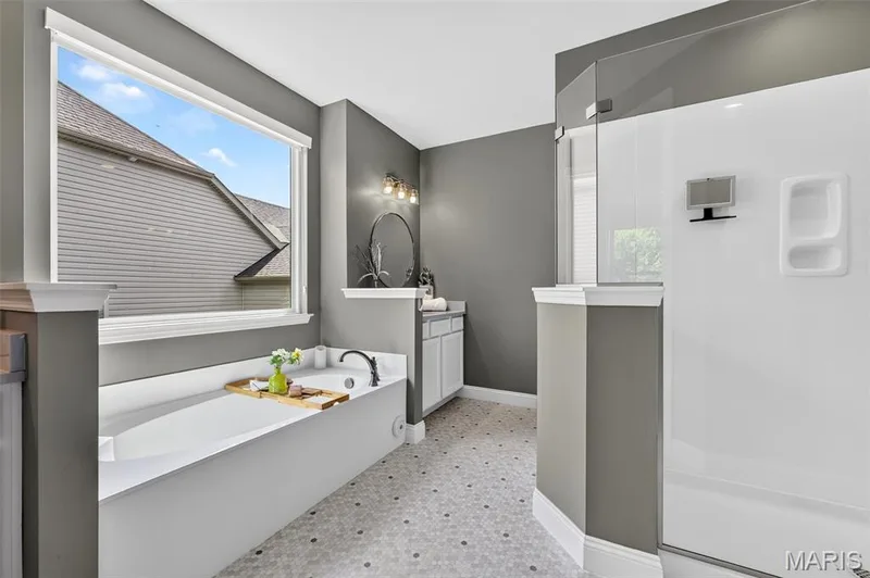 Soaking tub with black fixtures, walk-in shower with glass enclosure, single vanity with white cabinetry, wall-mounted light fixture, and hexagon tile flooring — 401 Hamlet Court, O'Fallon, MO 63367