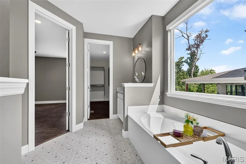 Soaking tub with a white countertop surround, large window, hexagon tile flooring, and a vanity with a round mirror — 401 Hamlet Court, O'Fallon, MO 63367