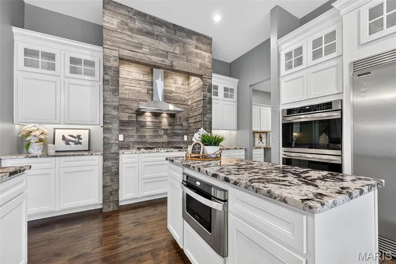Gourmet kitchen featuring white cabinetry, stone slab countertops, stainless steel appliances, a built-in microwave, and wood-finish flooring — 401 Hamlet Court, O'Fallon, MO 63367