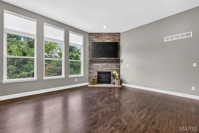 Wood-finish flooring, stone-clad fireplace with a wood mantel, and multiple large windows with roller shades — 401 Hamlet Court, O'Fallon, MO 63367