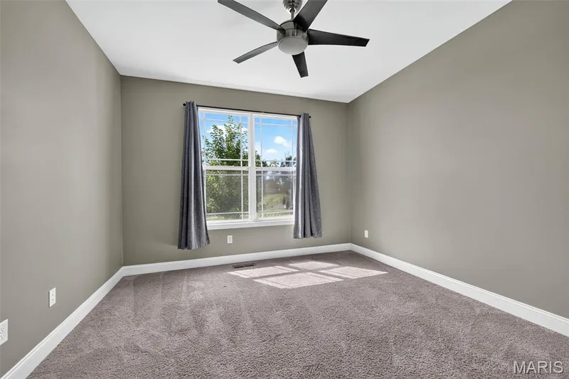 Ceiling fan with dark blades, large window with white trim, gray plush carpeting, neutral wall color, and white baseboards — 401 Hamlet Court, O'Fallon, MO 63367