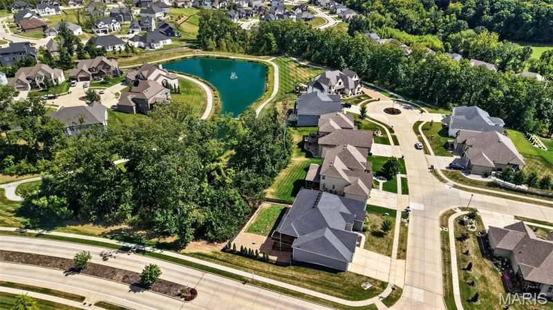 Aerial perspective showcasing a residential community with varied architectural styles, mature trees, and a central pond — 401 Hamlet Court, O'Fallon, MO 63367