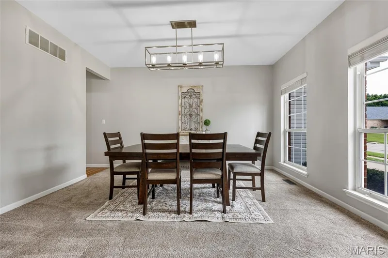 Modern design meets architectural charm. This carpeted room pairs tailored white-blinded windows and a minimalist rectangular chandelier with the soft curves of an open archway, all set against a calming backdrop of light gray walls. — 501 Oxborough Court, Cottleville, MO 63376