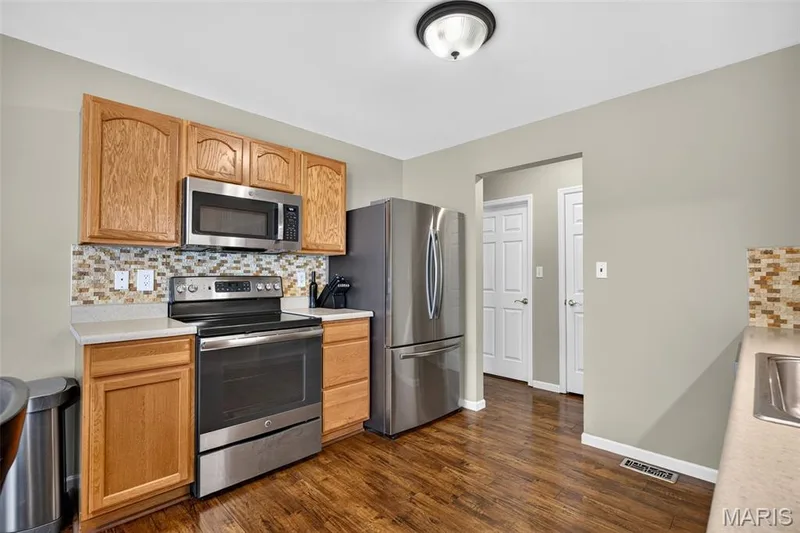 Kitchen featuring stainless steel appliances, wood-finish flooring, light wood cabinetry, stone-look countertops, and a mosaic tile backsplash — 30 Hunting Manor Drive, St Charles, MO 63303