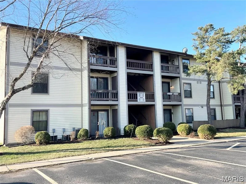 Multi-story building featuring light-colored siding with dark trim, private balconies/patios with wooden railings, and surrounding mature trees — 1430 Summergate Parkway #J, St Charles, MO 63303