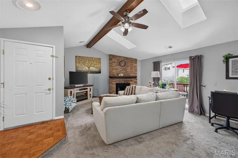 Living area featuring vaulted ceilings with exposed wood beams and skylights, a brick woodburning fireplace, and sliding glass door to outdoor space. — 974 Windstream Drive #38, St Peters, MO 63376