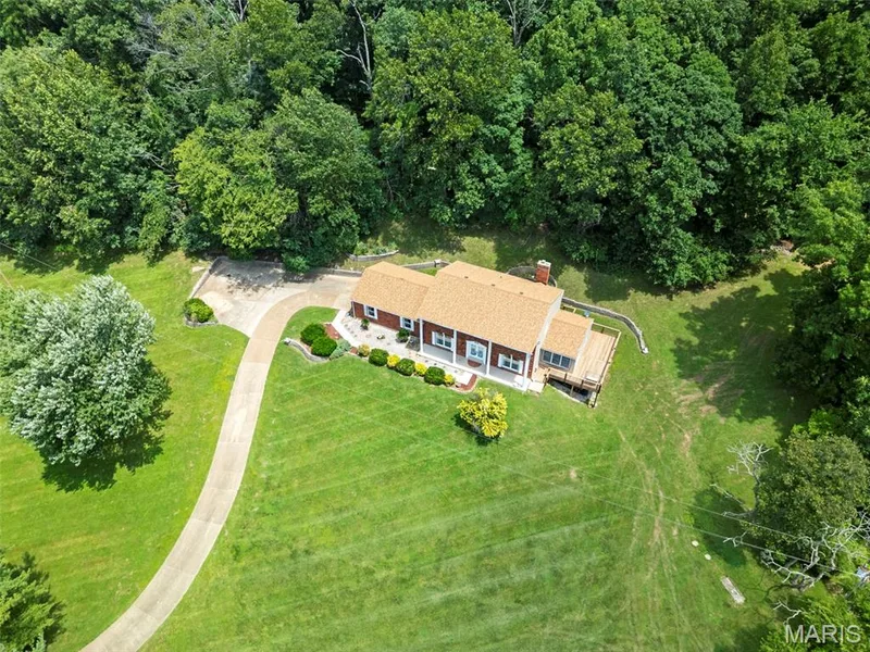 Red brick exterior with a light brown shingle roof, featuring a concrete driveway, expansive lawn, mature tree coverage, and an elevated wood deck — 3672 Hawks Rest Road, Wildwood, MO 63069