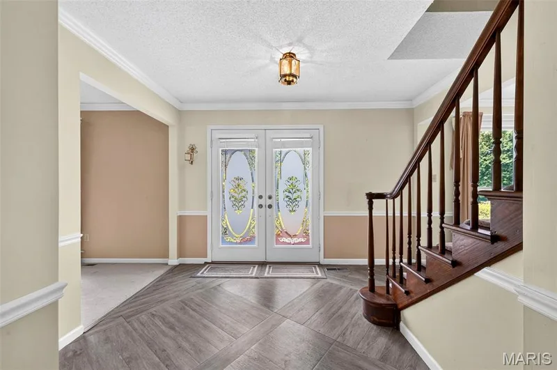 Entryway featuring decorative double doors, wood-finish flooring with a herringbone pattern, a dark wood staircase with balusters, crown molding, and a traditional ceiling light fixture — 3672 Hawks Rest Road, Wildwood, MO 63069