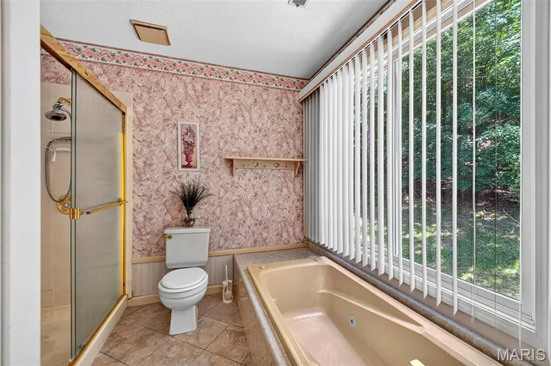 Bathroom featuring a glass-enclosed shower with gold-tone framing, a built-in tub, a large window with vertical blinds, and tile flooring — 3672 Hawks Rest Road, Wildwood, MO 63069