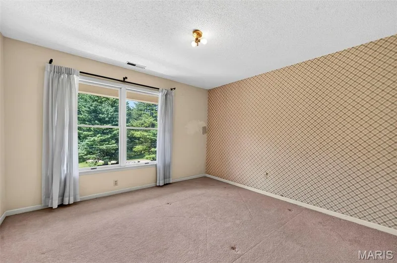 Room featuring a large double window overlooking extensive greenery, neutral wall color, patterned accent wall, light brown carpet, and a ceiling-mounted light fixture — 3672 Hawks Rest Road, Wildwood, MO 63069