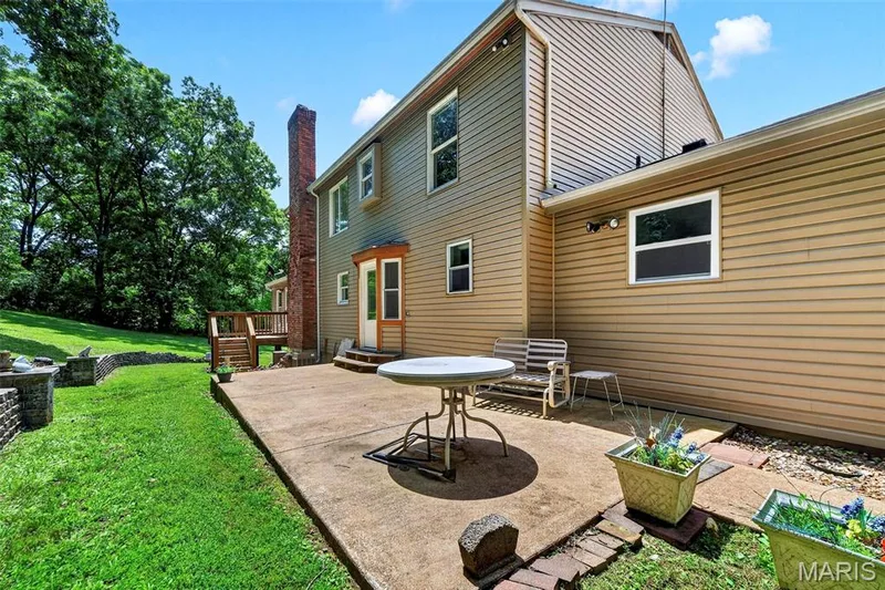 Rear elevation featuring tan siding, a concrete patio, and a tiered grassy area with stone retaining walls — 3672 Hawks Rest Road, Wildwood, MO 63069