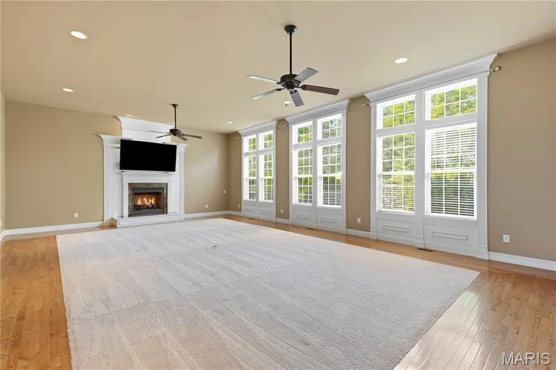Very large Great Room at 32x20 with an 11 foot ceiling, features a gas fireplace with stone surround and ornate mantelpiece, multiple large windows with stunning millwork, recessed lighting, and wood-finish flooring — 5500 Wooded Creek Drive, O'Fallon, MO 63304