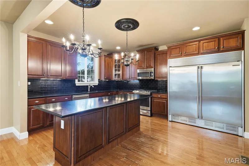 Kitchen featuring hardwood flooring, a 6 foot central island with granite countertop, extensive wood cabinetry, stainless steel appliances, and dark subway tile backsplash — 5500 Wooded Creek Drive, O'Fallon, MO 63304