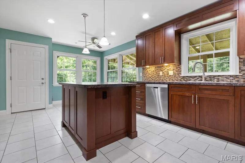 Kitchen featuring a central island, rich wood cabinetry, stainless steel dishwasher, mosaic tile backsplash, and light tile flooring — 44 Wilderness Lane, Defiance, MO 63341