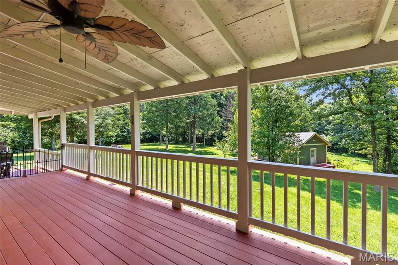 Spacious covered deck featuring red-stained wood flooring and white balusters, overlooking a private backyard with mature trees — 44 Wilderness Lane, Defiance, MO 63341