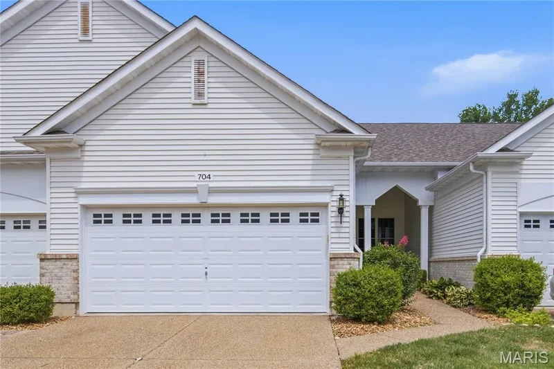 White vinyl siding exterior with an attached two-car garage featuring square window inserts — 704 Harrier Drive #V, Lake St. Louis, MO 63367