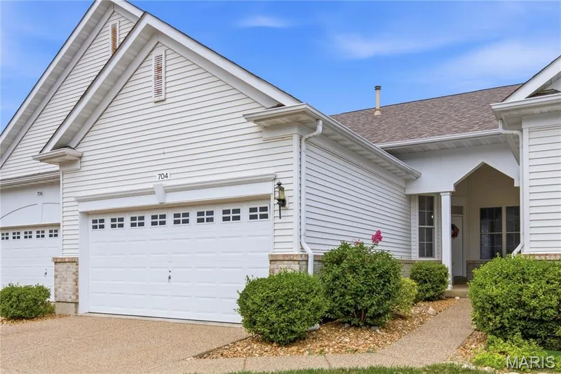 White siding exterior with an attached two-car garage featuring square window inserts — 704 Harrier Drive #V, Lake St. Louis, MO 63367