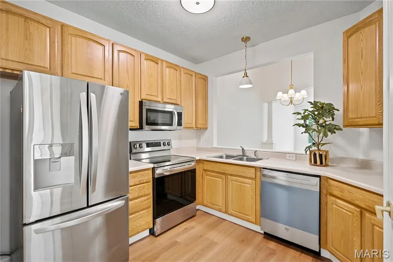Kitchen featuring wood-finish flooring, light wood cabinetry, stainless steel appliances, and a pass-through opening — 704 Harrier Drive #V, Lake St. Louis, MO 63367