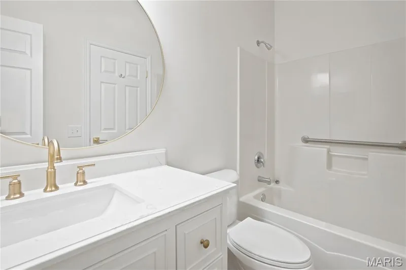 Bathroom featuring a white vanity with paneling, a white countertop, gold-finish fixtures, a round mirror, and a white tub with an integrated grab bar — 704 Harrier Drive #V, Lake St. Louis, MO 63367