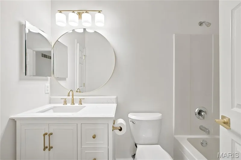 Bathroom vanity featuring a white shaker-style cabinet, a white countertop, a circular framed mirror, and a four-light fixture with brushed gold finishes — 704 Harrier Drive #V, Lake St. Louis, MO 63367