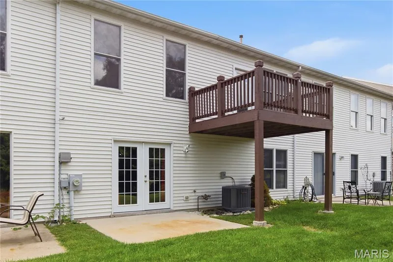 Rear exterior featuring white vinyl siding, a raised wood deck with dark stain, and a ground-level concrete patio — 704 Harrier Drive #V, Lake St. Louis, MO 63367