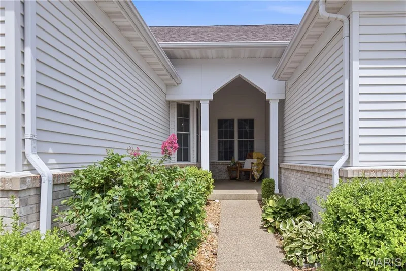 Inviting front entry featuring a covered porch with white columns, light siding, a brick facade, and a concrete walkway bordered by mature landscaping — 704 Harrier Drive #V, Lake St. Louis, MO 63367