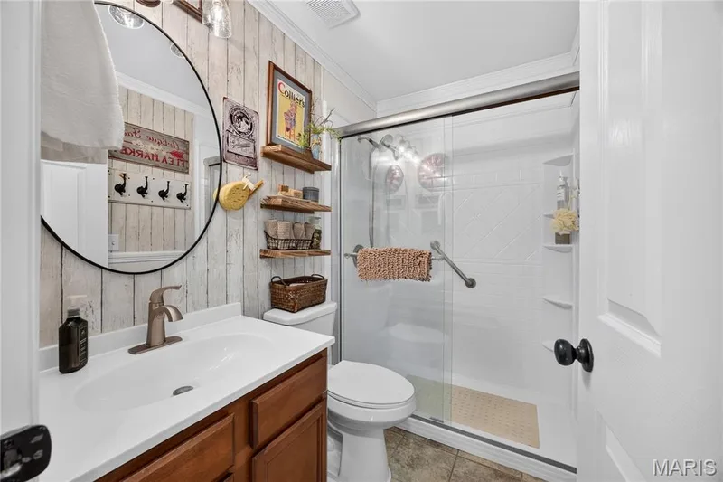 Bathroom featuring a walk-in shower with a glass enclosure, a vanity with a white countertop and wood-finish cabinetry, a round wall-mounted mirror, and plank-style wall paneling — 802 Barbara Place, O'Fallon, MO 63366