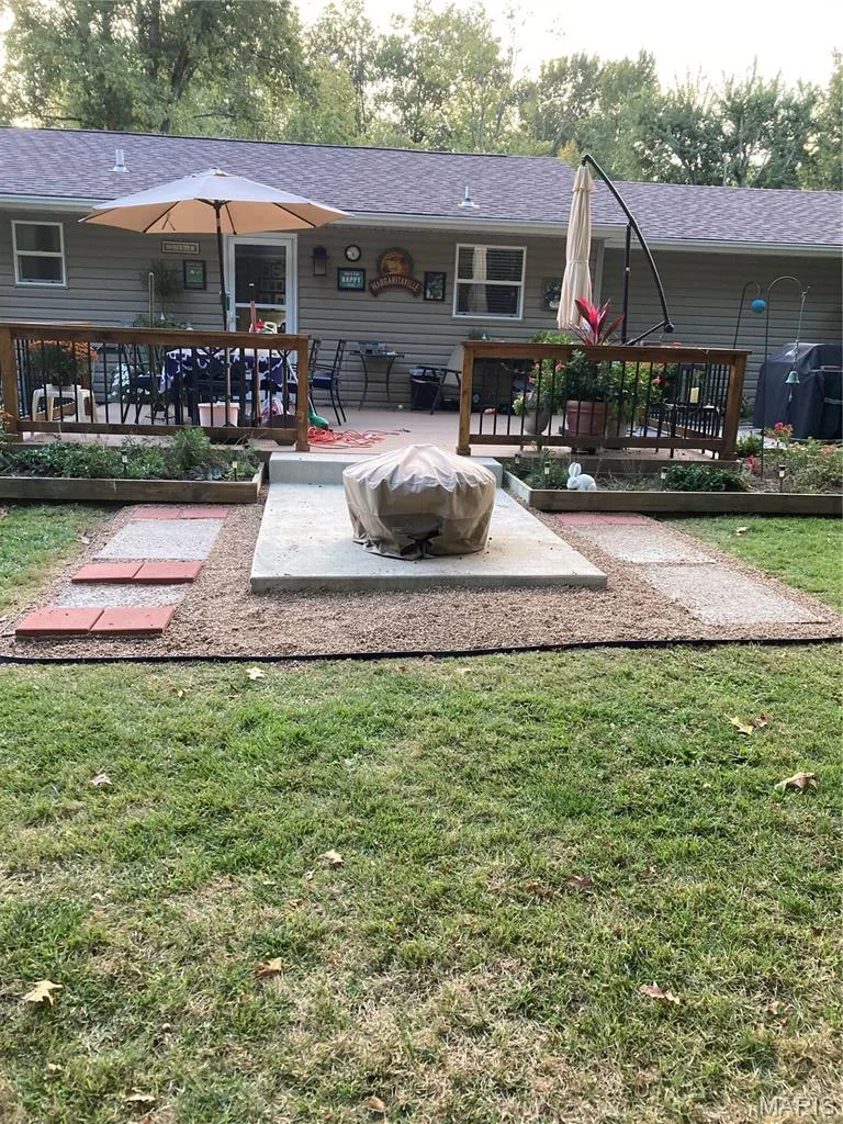 Expansive backyard featuring a concrete patio, wood deck with railing, and a central gravel-covered fire pit area — 802 Barbara Place, O'Fallon, MO 63366