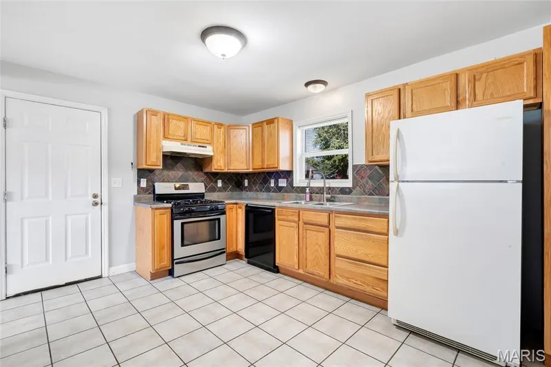 Kitchen featuring wood cabinetry, natural stone-look backsplash, stainless steel gas range, and ceramic tile flooring — 503 Eggering Drive, O'Fallon, MO 63366