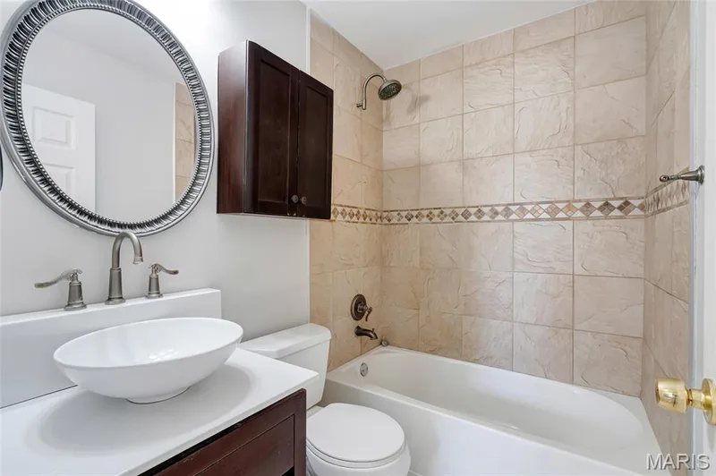 Bathroom featuring a white vessel sink, vanity with solid-surface countertop, dark wood-finish wall cabinet, and a tiled tub surround with decorative inlay — 503 Eggering Drive, O'Fallon, MO 63366