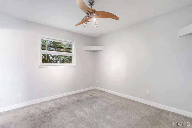 Neutral-toned interior featuring wall-to-wall carpeting, a multi-blade ceiling fan with integrated lighting, a single window, and a built-in corner shelf — 503 Eggering Drive, O'Fallon, MO 63366