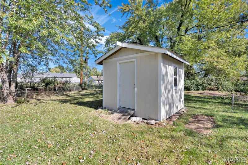 Exterior storage shed featuring light-toned vertical siding, a pitched roof, a single-entry door with a ramp, and mature trees on a grass lot — 503 Eggering Drive, O'Fallon, MO 63366