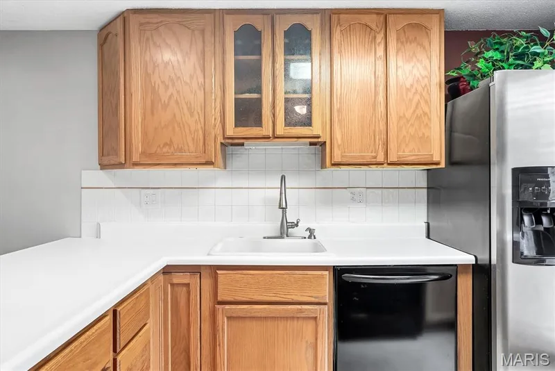 Kitchen featuring wood cabinetry, white countertops, a white tile backsplash, a stainless steel refrigerator, and a black dishwasher — 43 Breezy Knoll Lane, Lake St. Louis, MO 63367