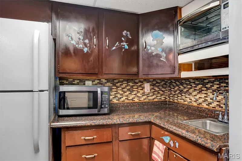 Compact kitchen area featuring wood-finish cabinetry, a mosaic tile backsplash, and a corner sink with a chrome faucet — 43 Breezy Knoll Lane, Lake St. Louis, MO 63367