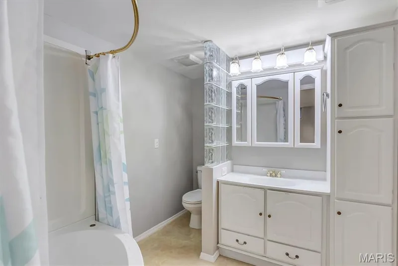 Bathroom featuring a glass block partition, white vanity with integrated sink, white storage cabinets, mirrored medicine cabinet, and a shower-tub combination — 43 Breezy Knoll Lane, Lake St. Louis, MO 63367