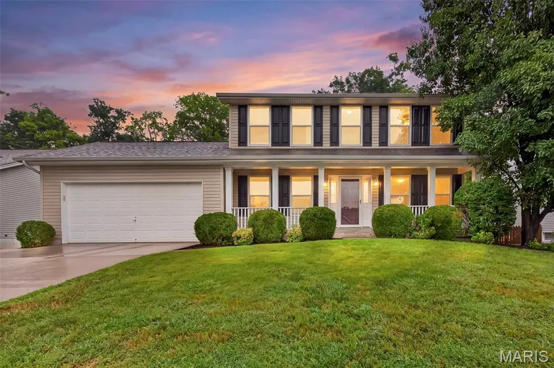 Two-story residence featuring light-colored siding, a covered front porch with white columns and railings, and a two-car garage with a concrete driveway — 4014 Treeshadow Drive, St Peters, MO 63376