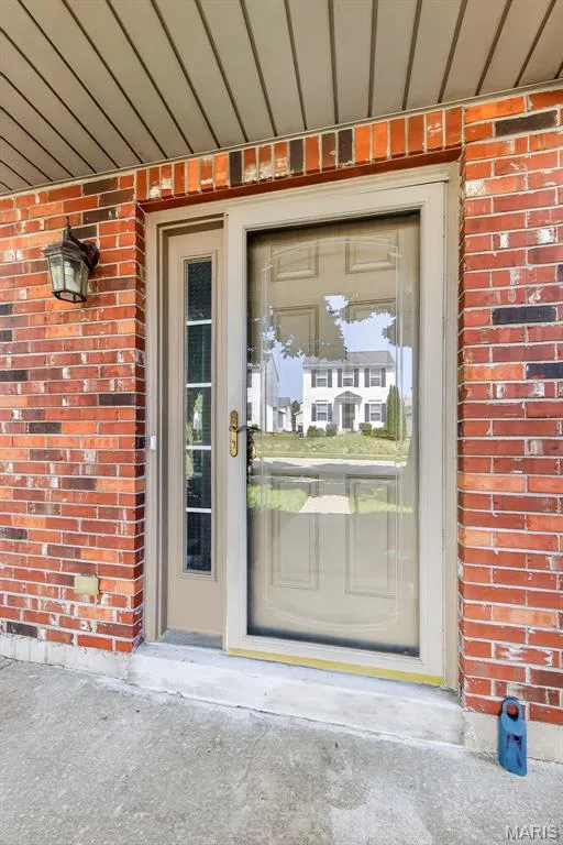 Brick exterior with a covered entryway, showcasing a tan front door with decorative glass panels and an adjacent full-view storm door — 916 Carpathian Drive, Lake St. Louis, MO 63367