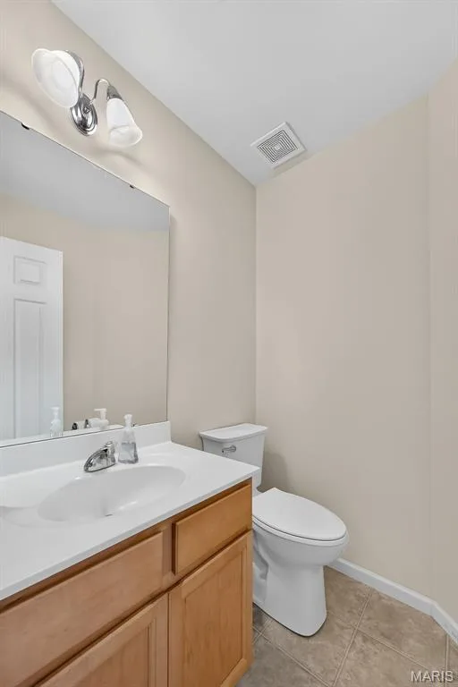Main floor 1/2 Bathroom featuring a wood-finish vanity with a white countertop and integrated sink, a wall-mounted mirror, and tile flooring — 916 Carpathian Drive, Lake St. Louis, MO 63367