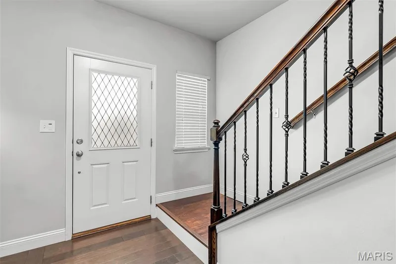 Welcoming foyer featuring a white six-panel entry door with diamond-pattern glass insert, hardwood flooring, and elegant staircase with dark wood handrail and decorative metal balusters — 225 Thorn Brook Drive, O'Fallon, MO 63366
