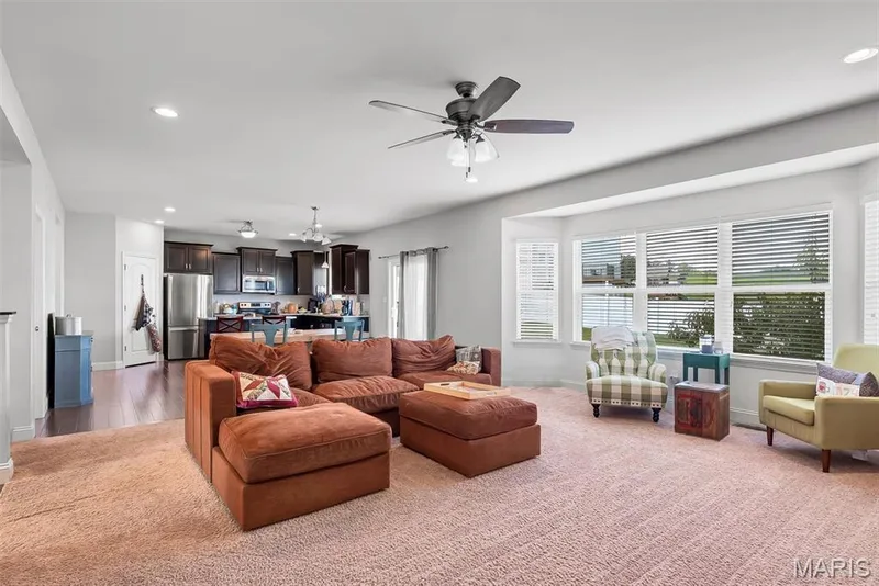 Open-concept living area featuring a spacious layout with plush upholstered sectional seating, neutral carpet flooring, and abundant natural light from multiple large windows with plantation shutters — 225 Thorn Brook Drive, O'Fallon, MO 63366
