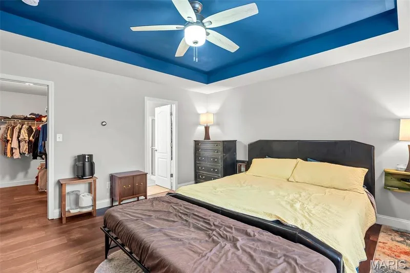 Spacious primary bedroom featuring a tray ceiling with contrasting blue finish and integrated ceiling fan, wood-finish flooring throughout, and a dark upholstered bed frame — 225 Thorn Brook Drive, O'Fallon, MO 63366