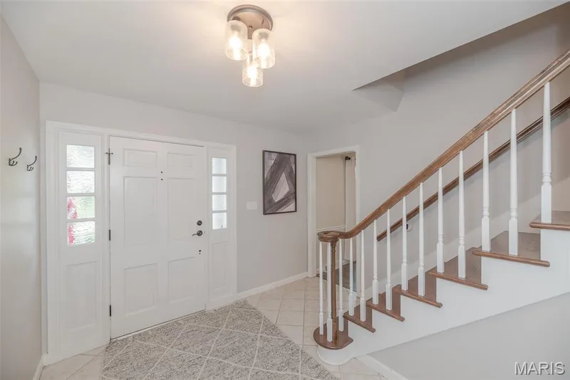 Bright foyer featuring a white six-panel entry door with sidelights, geometric tile flooring, a decorative ceiling fixture, and a wooden staircase with white spindles and a natural wood handrail leading to the upper level — 1526 Candish Lane, Chesterfield, MO 63017