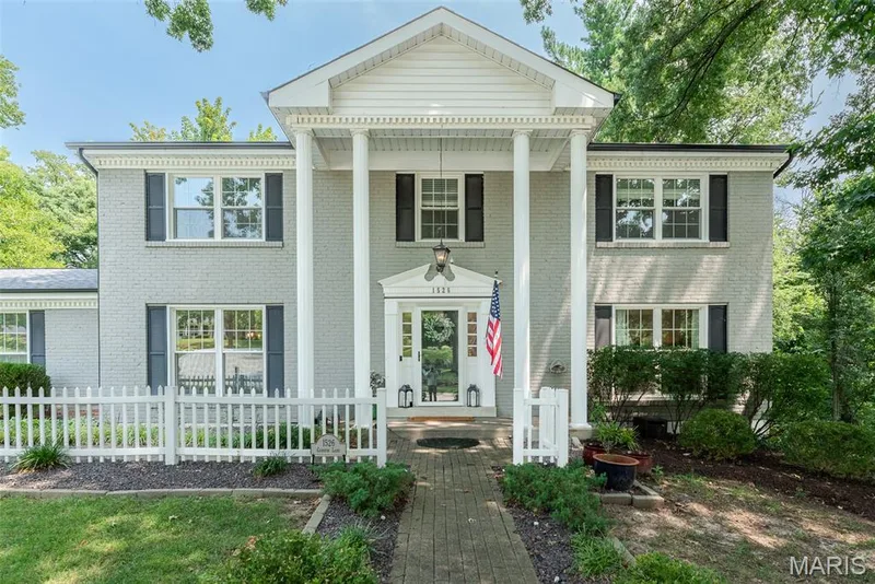 Traditional brick exterior with painted finish, featuring a prominent portico with white columns, a white picket fence, a brick pathway, and mature surrounding trees — 1526 Candish Lane, Chesterfield, MO 63017