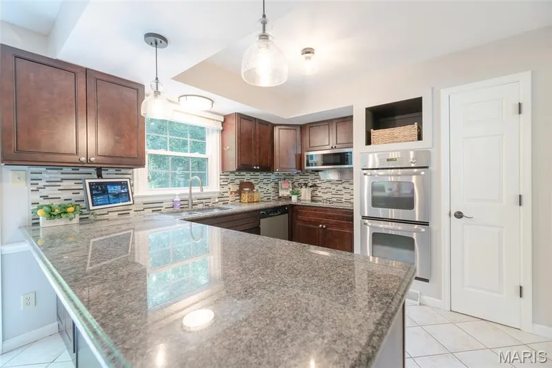 Elegant kitchen showcasing rich wood cabinetry, granite countertops with waterfall island edge, stainless steel appliances including double wall ovens, mosaic tile backsplash, pendant lighting, and tile flooring throughout — 1526 Candish Lane, Chesterfield, MO 63017