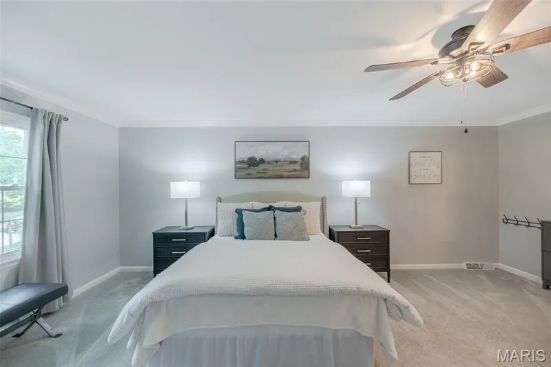 Spacious primary bedroom featuring dual nightstands with task lighting, wood-finish flooring, ceiling fan with integrated light fixture, and large windows with thermal-lined curtains — 1526 Candish Lane, Chesterfield, MO 63017