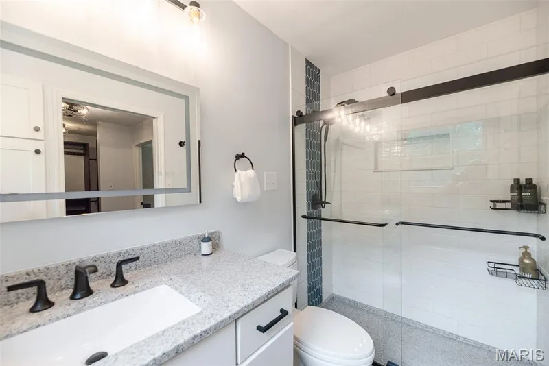 Modern bathroom featuring a vanity with a light-colored countertop and integrated sink, a large illuminated mirror, and a walk-in shower with a sliding glass door and subway tile surround — 1526 Candish Lane, Chesterfield, MO 63017