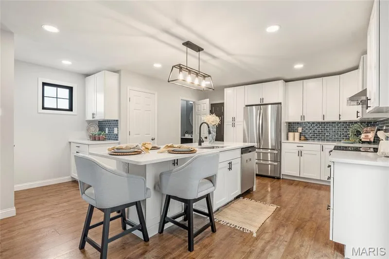 Bright kitchen featuring an island with integrated sink, white cabinetry, stainless steel appliances, a tile backsplash, and wood-finish flooring — 3429 Sublette Street, St Charles, MO 63301