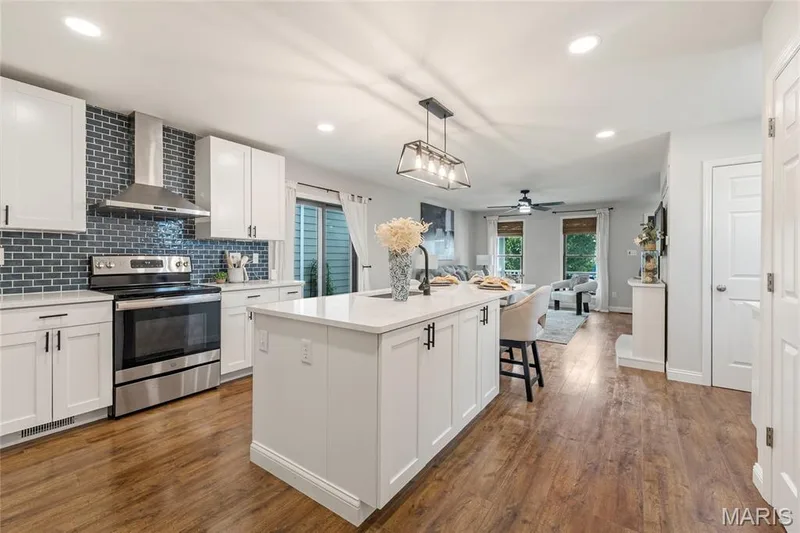 Modern kitchen featuring a large central island with a solid surface countertop, white shaker-style cabinetry with matte black hardware, a stainless steel range with matching hood, and wood-finish flooring — 3429 Sublette Street, St Charles, MO 63301